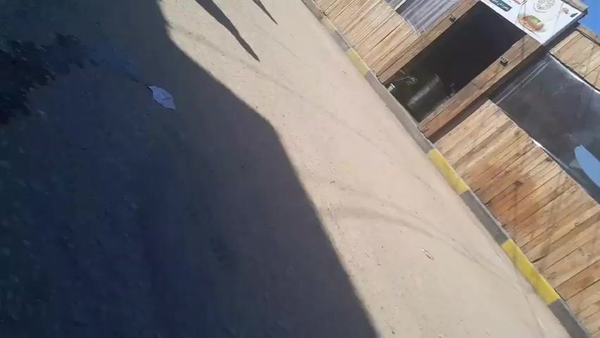 The video shows a street scene with a building made of wood. There are palm trees in the background and a blue sky. A sign with Arabic writing is attached to the building. Shadows of people can be seen on the ground. There are a few scattered items on the sidewalk. The camera angle is tilted.