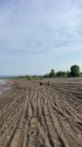 Two young males, one appearing older than the other, are running away from the camera on a dirt path. The path is marked with tire tracks. The environment appears to be a sandy, open area with some vegetation in the distance and a body of water to the left. The sky is blue with scattered clouds. Both males are shirtless, wearing shorts. They are running side by side, with the older male slightly ahead.