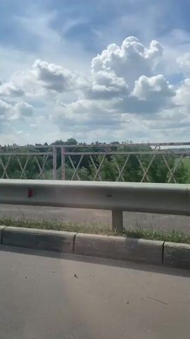 The video pans across a bridge on a bright, cloudy day. The foreground shows the asphalt road surface, a concrete curb, and a metal guardrail. Behind the guardrail, a red metal railing with a diamond pattern is visible. Beyond the railing, a lush green landscape with trees stretches into the distance. The sky is filled with large, fluffy white clouds against a bright blue backdrop, suggesting it is daytime, likely midday or early afternoon. The weather appears pleasant, with the sun shining through the clouds. The location is identified as Zaraysk, Russia. There are no people visible in the video, and no text is present. The overall mood is peaceful and scenic, capturing a travel or transit moment.