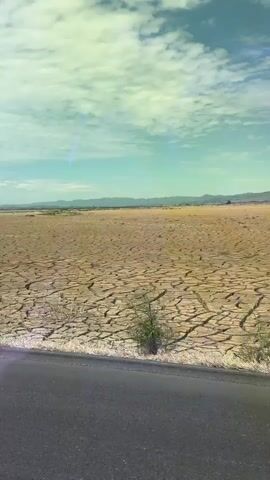 A video shows a cracked, dry landscape under a cloudy sky. The ground is parched and resembles dried mud, with large fissures crisscrossing the surface. Sparse, dry vegetation dots the edges of the land. In the foreground, a dark asphalt road is visible. In the distance, a range of hills or mountains can be seen under the hazy sky. The scene appears to depict an area experiencing drought.