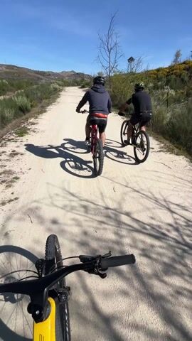 Three people are biking on a dirt road. The person recording is riding a yellow "STAKE" mountain bike. The other two bikers are riding ahead. The biker in the back is wearing a navy blue hooded sweatshirt and red shorts. The biker in the middle is wearing a black shirt and shorts. The road is surrounded by trees and shrubs. The sky is blue. At one point, the recording biker takes a selfie, giving a thumbs up while wearing an orange and black biking shirt.