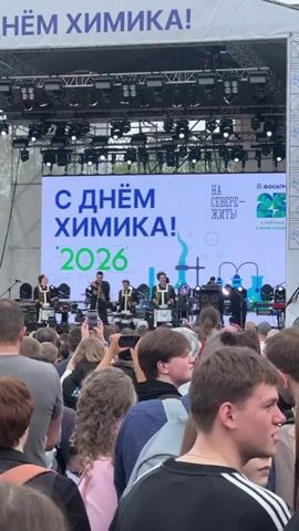 The video shows a large crowd watching a band perform on an outdoor stage. The band members are dressed in matching black outfits with gold trim and are playing drums. Behind the band is a large screen that says "С Днем Химика! 2026" (Happy Chemist's Day! 2026). The event appears to be sponsored by "ФосАгро" (PhosAgro). Many in the crowd are recording the performance on their phones.