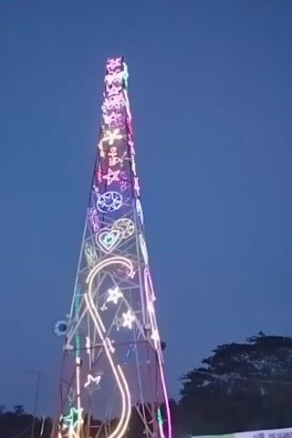 The video captures an outdoor scene at dusk or early night, featuring a tall, slender, tower-like structure brightly illuminated with colorful LED lights. The lights form various decorative shapes, including stars, hearts, anchors, and what appears to be a large stylized 'S' or '8' at the base, primarily in pink, purple, blue, green, and white. To the left, a portion of a Ferris wheel, also adorned with vibrant lights, is visible against the dark blue sky. Trees are present in the background, flanking the structures. The illuminated attractions suggest a festive event, likely an amusement park or a fair in Charfasson, Bangladesh. A sign with partially visible text in Bengali script, reading "দুই কুম" (Dui Kum) is seen at the bottom right. The atmosphere conveyed is lively and celebratory.