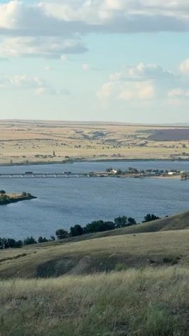 A landscape view from a grassy hill overlooks a wide, blue river. A bridge with several vehicles crosses the river in the mid-ground. On the far side of the river, there are fields of dry, golden grass and some scattered trees. The sky is a pale blue with scattered white clouds. The overall impression is of a rural, open landscape under a bright sky.