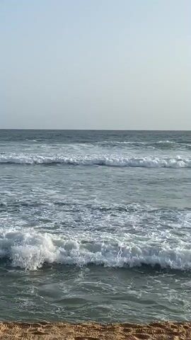 The video shows a serene beach scene with gentle waves lapping against the shore. The sky is a pale blue, with the sun casting a warm glow over the water. In the foreground, a sandy beach stretches out, with a closed beach umbrella and a wooden pole adding a touch of man-made presence to the natural landscape. A few distant rocks break the horizon, adding depth to the scene. The overall mood is tranquil and peaceful, inviting viewers to imagine themselves relaxing by the sea.