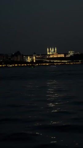 The video shows a dark body of water with a city skyline lit up in the background. A small boat with red lights is moving across the water.