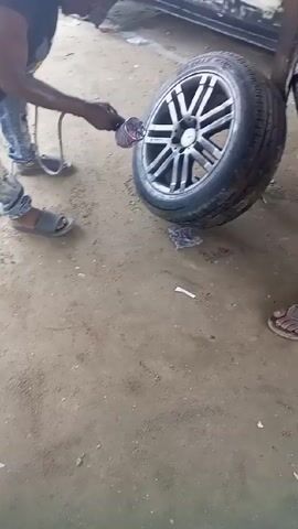 A man is painting a tire with an airbrush. He is wearing a black shirt, jeans, and sandals. The tire is leaning against a wooden post. Another man is sitting nearby. The ground is dirt. There is a car in the background.