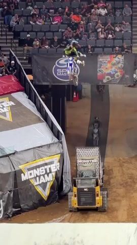 A motocross rider is performing a jump over a small construction vehicle. The rider is in mid-air, on a white and blue dirt bike, with their body angled forward. The vehicle, a yellow compact loader, is positioned below and to the right of the rider. The background consists of stadium seating filled with spectators. Another dirt bike is visible further back on a ramp. The event appears to be Monster Jam, as indicated by branding on the ramp and the vehicle.