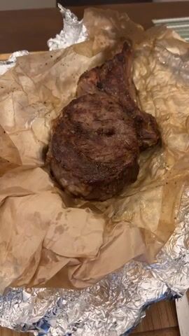A bone-in steak, wrapped in parchment paper and aluminum foil, is presented on a wooden surface. The steak is seared and seasoned, with a thick cut of meat and a bone protruding from one side. The parchment paper is slightly greasy, indicating that the steak has recently been cooked. The aluminum foil is crumpled around the base of the steak, providing a makeshift serving dish. The lighting is warm and inviting, suggesting a home-cooked meal.