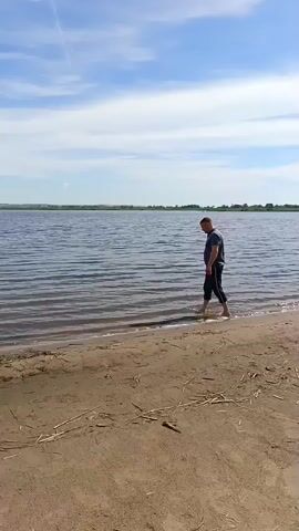A man walks into the water from a sandy shore. The man is wearing a dark t-shirt and dark pants. He is walking into a large body of water with gentle waves. The sky is blue with scattered clouds. The background shows a distant tree line.