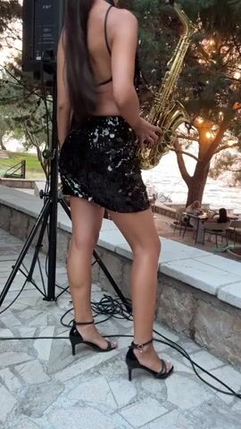 A woman with long dark hair tied in a ponytail stands on a stone-paved patio, holding a golden saxophone. She is wearing a black sequined mini-skirt and a black bikini top, with black strappy heeled sandals. Her left hand is on the saxophone, and she appears to be playing it. To her left, a black speaker on a stand is visible. The scene is outdoors, with greenery and trees behind her, and the sea or a large body of water in the background. A few tables and chairs are set up in the background, suggesting a restaurant or outdoor seating area. The sun appears to be setting, casting a warm glow over the scene and creating long shadows, indicating it is late afternoon or early evening. The woman's stance and focus on the saxophone suggest she is performing. The overall atmosphere appears relaxed and pleasant, possibly a live music performance at a coastal venue. Text visible on the speaker reads "FBT MAXX 26". The location context is Pečurice, Montenegro.