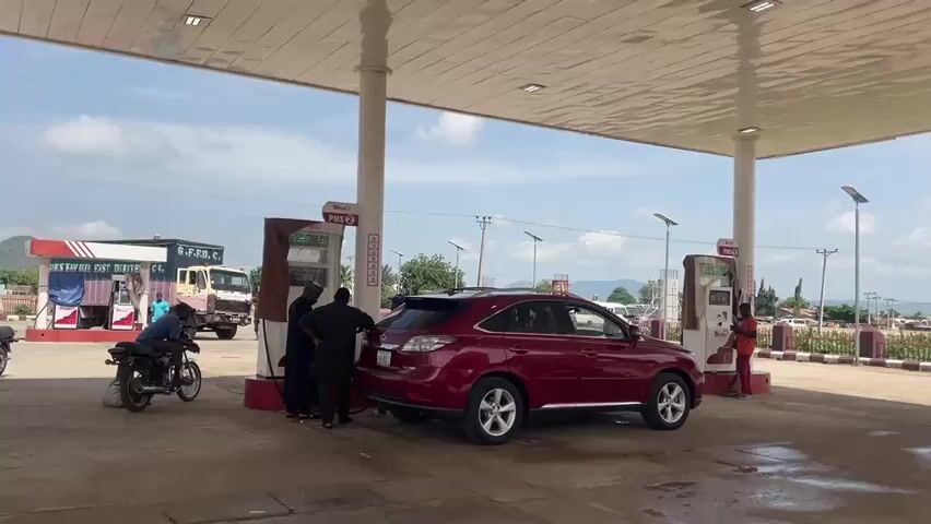 The media file captures a scene at a gas station in Pasere, Nigeria. The setting appears to be outdoors, under the shelter of a large canopy, with a bright, sunny sky and scattered clouds, suggesting it is daytime. The ground is paved with concrete, and there are several gas pumps visible.

The primary activity is refueling vehicles. A red SUV is parked at one of the gas pumps, with people attending to it, presumably filling its tank. To the left, a motorcycle is also parked, and a person is seated on it. Further to the left, a truck is visible, and another person appears to be standing near it. In the background, there are buildings, a cell tower, and some vegetation, with hills or mountains in the distance. The overall atmosphere seems ordinary, with no immediate signs of distress or heightened emotion.

Visible text includes branding on the gas pumps, such as "Shafi" and "PMS," which is a common term for Premium Motor Spirit (gasoline) in Nigeria. There is also text on the side of the truck, which appears to read "B.S.R.C. EASTERN DISTRIBUTOR." The presence of multiple people, vehicles, and the gas station infrastructure all point to a typical operational day at this location. The lighting and shadows suggest it is likely mid-morning or afternoon.