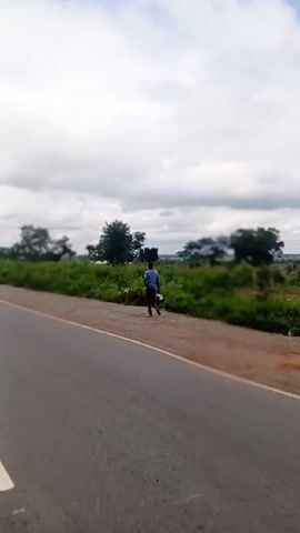 The video shows a view from a vehicle moving along a paved road. On one side of the road is green vegetation and trees. The sky is cloudy. A person on a motorcycle passes in the same direction as the camera vehicle.  The road has a yellow line on the shoulder and a white dashed line dividing the lanes.