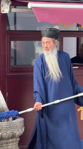 Here is a description of the video.

A man wearing a blue robe and a black cap stands in front of a maroon-colored building. He has long, white facial hair. He holds a mop and cleans a stone structure. The mop has a blue, microfiber cleaning head attached to a white pole. The stone structure is decorated with a dragon head and is positioned in front of a maroon building with a red awning above. The man uses the mop to clean the stone structure, immersing it in water and then scrubbing the surface. The camera is positioned at eye-level and remains relatively still throughout the brief video.