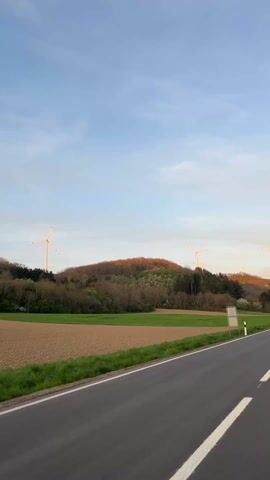 The camera records a landscape on a clear day. A paved road runs alongside a field, bordered by a green strip of grass. Beyond the field, a tree-covered hill rises, topped with multiple wind turbines. The turbines appear to be illuminated by the setting sun. The sky is a mix of blue and pale white.