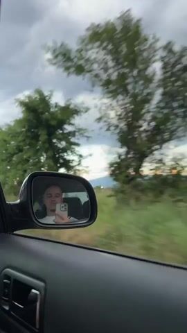 The video captures a view from inside a moving car, looking out the passenger side window. In the car's side mirror, a young man is visible, holding a light-colored smartphone. He appears to be recording or taking a selfie.

Outside the window, a green, somewhat blurred landscape rushes by, indicating the vehicle is in motion. Tall trees with lush green foliage line the side of the road, along with grass and other vegetation. Overhead, power lines stretch across a cloudy, overcast sky.

As the car moves, two road signs become visible: a yellow diamond-shaped sign, typically indicating a priority road in European traffic, and a blue circular sign with white arrows pointing right and curving left, which often signifies a mandatory direction of travel or a roundabout ahead. In the distance, a hazy horizon can be seen, possibly suggesting late afternoon or early evening light filtering through the clouds. The setting appears to be a rural or semi-rural area near Voluyak, Bulgaria.