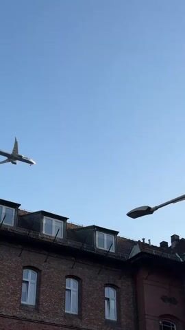 The video captures an airplane flying low over a brick building with a sloped roof. A street lamp is positioned in the foreground, partially obscuring the view. The sky is clear and blue. The airplane appears to be a commercial jet with its landing gear down. The building has multiple windows and dormer windows on the roof. The video is shot from a low angle looking upwards.