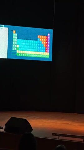 A man is on stage speaking into a microphone in an auditorium or theater. He is wearing a light-colored jacket, a dark shirt, and brown trousers, and appears to be giving a presentation or lecture. A large screen behind him displays a colorful periodic table of elements. The stage is dimly lit, with a spotlight on the speaker and the illuminated screen. The foreground shows the heads of audience members. A large black speaker is visible on the stage floor to the left. The man gestures with his hands as he speaks. The overall setting suggests an educational or public speaking event.