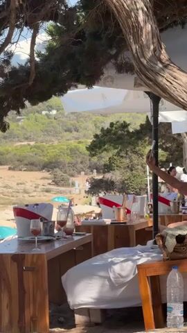 The video shows a view from a shaded outdoor seating area, likely a beach club or restaurant, looking out towards a beach and a hillside. In the foreground, there are wooden tables with white cushions on benches. On one table, there are two glasses of red liquid, two white buckets with red trim, and what appears to be a bottle. In the background, there are umbrellas and people on a sandy beach. A person's arm and hand holding a phone is visible on the right side of the frame, seemingly taking a photo. The environment appears to be sunny and arid with sparse vegetation on the hillside.