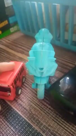 A human hand is seen from the left of the frame nudging a red toy bus forward. The bus has a cartoonish face printed on its front, with large eyes and a smiling mouth. It also has the number 1088 printed on its side. To the right of the bus is a turquoise toy motorcycle. The motorcycle is positioned upright and faces away from the camera. Its design suggests it is a sports bike. Part of a black object is visible on the far right edge of the frame. The toys are placed on a surface that appears to be a wooden table with a striped pattern. In the background, a light blue plastic basket is visible, containing some items that are out of focus. The lighting suggests an indoor environment.