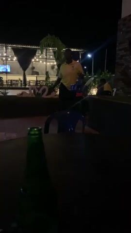 A person wearing a yellow shirt and black pants stands holding a blue bottle carrier with several bottles in it. They are in an outdoor setting at night, possibly a restaurant or bar, with tables and chairs visible. The person is walking towards a table in the foreground where a green bottle is placed. Lights are strung up in the background, and there are palm trees. The person appears to be serving drinks.