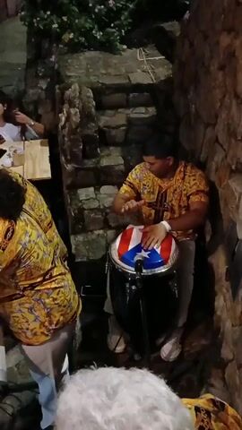 Here is a description of the video:
A musician in a vibrant patterned shirt plays a conga drum. The drum's head is decorated with the Puerto Rican flag. The musician sits in a small, stone-walled space with steps behind. A microphone sits in front of the drum. Other musicians with matching yellow shirts stand nearby. The lighting is dim.