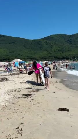 The video shows a sandy beach on a clear, sunny day with a blue sky and green, forested hills in the background. Several people are visible on the beach. In the foreground, two individuals are walking away from the camera towards the water, one carrying a pink paddleboard and the other carrying a white paddleboard. Further down the beach, there are more people, some sitting on chairs under umbrellas, and others in the shallow water. The ocean has gentle waves and is a turquoise color. There is some seaweed on the sand in the foreground.