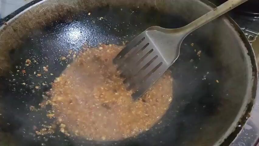 In a close-up shot, a person uses a slotted spatula to stir a bubbling mixture in a dark, well-used wok. The mixture appears to consist of finely chopped ingredients, possibly garlic and chili peppers, combined with a liquid base. The wok shows signs of previous use, with some discoloration and residue on the interior surface. The scene is brightly lit, highlighting the textures and colors of the cooking process.