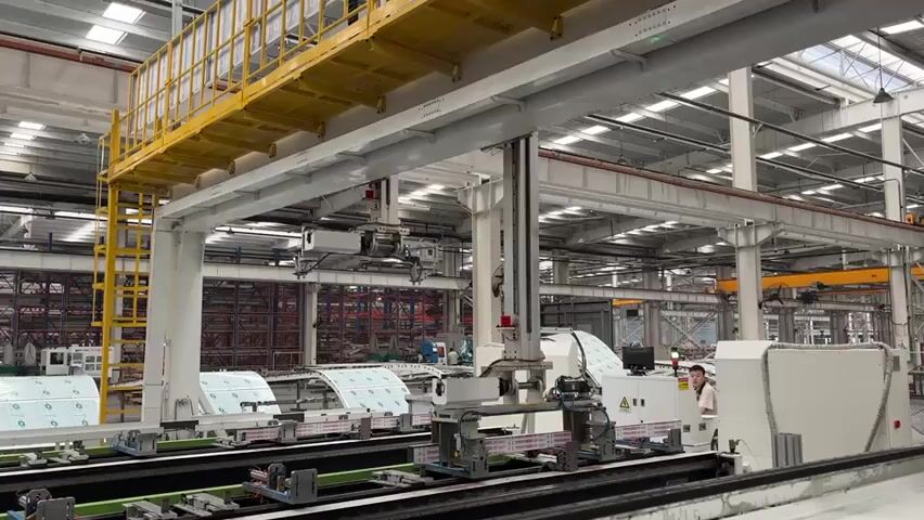 The video shows the interior of a large, brightly lit industrial facility. The focus is on a white, automated manufacturing machine with several arms suspended from overhead beams, moving along a track. The machine is processing what appear to be large sheets of material with a green and white pattern. In the background, there is a yellow maintenance platform with a ladder and more industrial equipment. A person wearing a white shirt is seen near the machine, possibly monitoring the operation. The environment is characterized by a high ceiling, numerous steel beams, and other industrial machinery, indicative of a large-scale production or manufacturing setting.