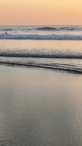 The video shows a close-up, eye-level shot of a beach at sunset. The waves are gentle, rolling in towards the shore. The sand is wet and reflective, mirroring the soft orange and pink hues of the sky. The sun is setting on the horizon, casting a warm glow over the entire scene. There are no people or objects visible in the video.
