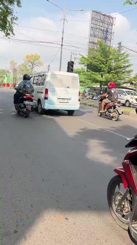 The video shows a man riding a red and black scooter on a paved road. He is wearing a blue t-shirt, white shorts, and a white helmet with blue and red accents. His right foot is visible wearing a flip-flop. The scooter has a license plate that reads W 5360 DB. In the background, a white van with blue lettering is visible, and another scooter with a rider is in front of the man. Traffic lights are visible, and trees line the side of the road. The sky is clear and blue, indicating a daytime scene. The overall environment appears to be an urban or suburban street in a tropical or subtropical region, likely Asia, given the type of vehicles and streetscape.