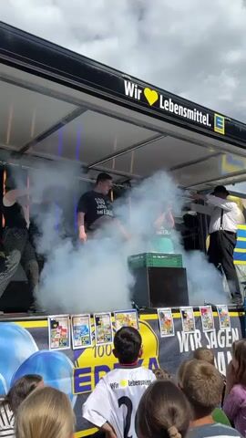A group of people are on a stage, possibly a parade float, with a black banner overhead that reads "Wir Lebensmittel. EDEKA." There is a lot of smoke in the air, obscuring some of the people. In the foreground, a crowd of children are watching. Some of the children are wearing white t-shirts with numbers on the back. There are also some balloons in the foreground. Several people are on the stage, wearing dark t-shirts with some text and one person in a white shirt and tie. There are also some green crates and what appears to be a black box on the stage. There are also some items displayed on the front of the stage, possibly flyers or merchandise. The sky above is cloudy.
