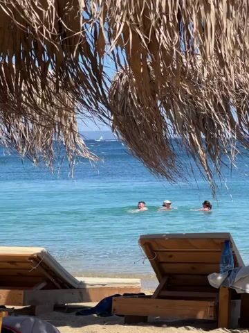 The video shows a beach scene with palm frond umbrellas and lounge chairs in the foreground. Three people are visible in the shallow, turquoise water, swimming. One person appears to be wearing a white hat. The water is calm and clear, and the sky is bright blue, suggesting a sunny day. A plastic water bottle is on one of the lounge chairs.