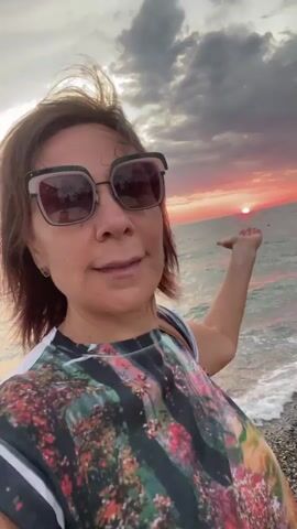 A woman stands on a beach at sunset. She is wearing large sunglasses and a colorful t-shirt. The sun is low in the sky, casting a pinkish glow over the clouds and the ocean. The woman has her right arm extended, with her palm facing upwards, as if to catch the sunlight. Waves are gently lapping at the shore.