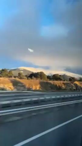 Here is a forensic description of the scene in the video:

The video shows a landscape view from a moving vehicle, likely a car or bus, traveling on a highway. The camera's perspective is through a window, which reflects a sign and the silhouette of a person. The primary focus is on the scenery outside.

To the right of the highway, there is a mountainous terrain covered with trees and patches of exposed earth. The mountaintops are covered in snow. The sky above is a mix of blue and dark, cloudy gray. The lighting suggests it is daytime, with some areas illuminated by sunlight.