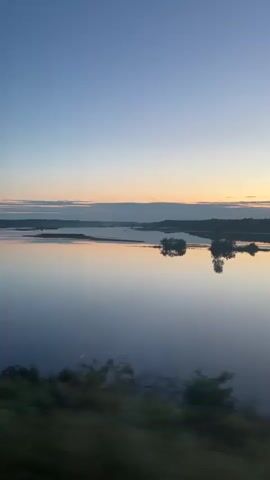 The video shows a view of a calm body of water at sunrise or sunset. The sky is a gradient of blue and orange, with wispy clouds. The water reflects the sky and the distant treeline, creating a mirror image. There are a few trees visible along the bank in the foreground, and the camera appears to be moving, causing a slight blur.