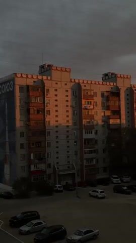The video shows a residential building during sunset. The building is made of concrete and has balconies. The sun is setting, casting a warm orange glow on the building. There are several cars parked in the courtyard. The sky is cloudy and dark.