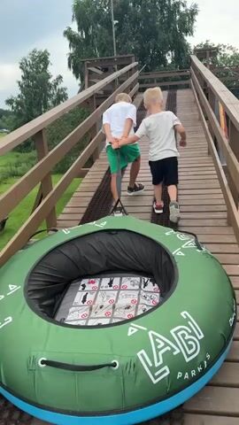 Two young boys are walking up a wooden ramp with a green and blue inflatable tube. The boy in front is wearing a white t-shirt and black shorts, while the boy behind him is wearing a white t-shirt and green shorts. They are holding onto a rope attached to the tube. The ramp leads to a slope with a green mesh surface and red trim. Trees and grass are visible in the background, along with a wooden structure at the top of the slope.