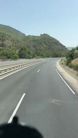 The video shows a point-of-view shot from a vehicle traveling on a highway. The highway has multiple lanes separated by guardrails and white lines. The surrounding environment is mountainous and covered with green trees. The sky is clear and blue, indicating a sunny day. The road ahead curves to the right. Lane markings on the road indicate a split in the lanes, with arrows showing that traffic can go straight or veer off to the right. A blue road sign is visible on the right side of the road. The camera is positioned low, likely from the dashboard of the vehicle, and the top of a black object, possibly a car accessory, is visible at the bottom of the frame. The overall scene depicts a scenic drive through a hilly landscape on a clear day.