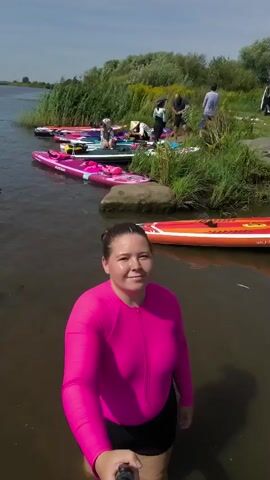 A woman in a pink rash guard and black shorts is holding a selfie stick in the water. Behind her, several stand-up paddleboards are lined up on the shore. A group of people are standing on the grass near the paddleboards, and a white van is parked in the background. The scene appears to be a recreational outing by a body of water on a sunny day.