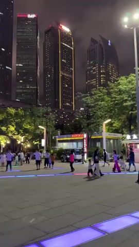 The video shows a bustling nighttime scene in Tianhe District, China. A group of people are walking across a paved plaza. The plaza is illuminated by a series of colorful, pulsing LED lights embedded in the ground, creating a vibrant and dynamic atmosphere. Towering skyscrapers, adorned with a multitude of lights, dominate the background, suggesting a modern urban environment. Some of these buildings display dynamic light patterns, with one appearing to have red and pink lights creating a large graphic on its facade. The sky is dark and cloudy, typical of a night in a city. The overall impression is one of lively urban activity and modern city lights, with people enjoying the evening in a well-lit public space. There is a small white vehicle, possibly an electric cart, visible in the middle ground. The camera movement is smooth, capturing the wide expanse of the plaza and the impressive cityscape.