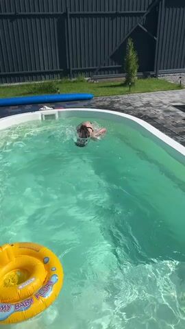 A child is swimming in a pool. The child is wearing a swimsuit and goggles. There is a yellow inflatable ring in the pool. The child is kicking their legs and splashing water. The child is looking at the camera. The pool is surrounded by a fence and a patio. There is a blue inflatable tube in the background.