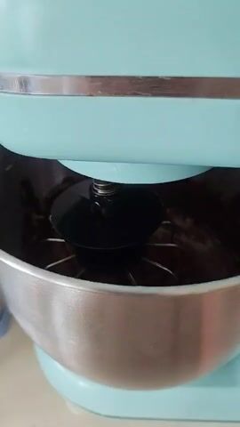 A close-up shot shows a whisk attachment on a stand mixer, coated in thick, dark chocolate batter. The whisk is in a stainless steel bowl, also containing the chocolate mixture. The mixer head is a light blue color. The batter appears smooth and glossy.