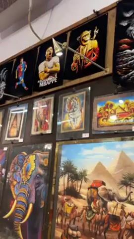 The video shows a display of framed and unframed artwork for sale. The display includes artwork with images of athletes, animals, pyramids, and cities. The artwork includes both paintings and tapestries. A person holds up a phone to take a picture of the artwork. The environment appears to be a market or store.