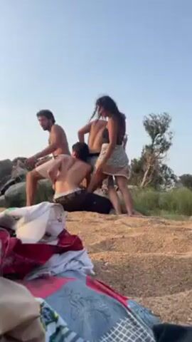 Four individuals stand on a sandy hill. Two men are shirtless, one man is wearing dark pants and the other is wearing light shorts. The other male is out of view. A woman wearing a black top and a printed wrap-around skirt also stands there. They are grouped together, interacting with one another. A pile of clothing, including a white baseball cap, lies in the foreground. In the background, there are rocks, green grass, and a tree. The sky is clear and blue.