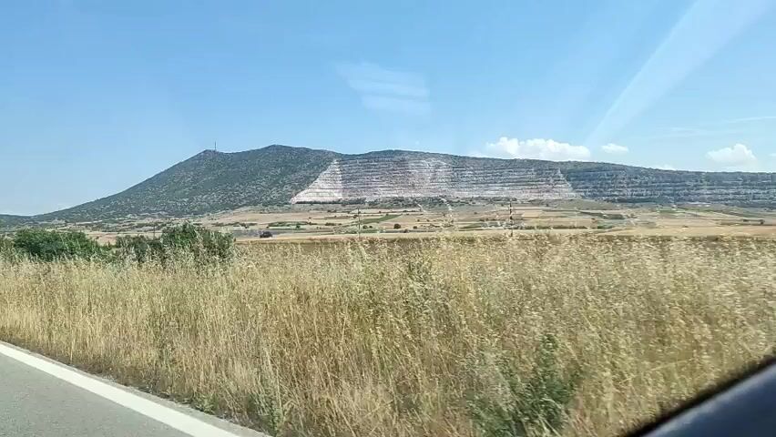 The video shows a driver's perspective from inside a moving vehicle, looking out the front passenger window. The scene outside depicts a sunny day with a clear blue sky and some white clouds. In the distance, a large, dark green mountain with a communication tower on its peak is visible. To the right of the mountain, a substantial white, terraced structure, possibly a quarry or mine, is carved into a hillside. In the mid-ground, a flat, dry, yellowish-brown field extends across the landscape, dotted with occasional small green bushes and trees. A power line with utility poles runs horizontally across the field. In the foreground, a strip of dry, tall, golden-brown grass lines the edge of the road, which has a white barrier visible on the left side. As the vehicle moves, the foreground elements, including the grass and bushes, pass by quickly.