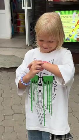 A young girl with blonde hair and a white t-shirt featuring a wolf graphic with green dripping sunglasses is looking at her wrist. She is wearing a beaded bracelet on her left wrist and a red and blue band on her right wrist. Another child, seen from behind, with long blonde hair tied in ponytails, approaches the first girl. The second child points at the first girl's wrist, and the first girl smiles. The second child then touches the first girl's bracelet. The environment appears to be outdoors, possibly near a store with displays visible in the background.