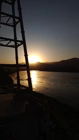 The video opens with a view of a wide river at sunset. A tall metal structure is on the left side of the frame. The sun is low on the horizon, casting a bright reflection on the water. In the distance, mountains and a city skyline are visible. Several cable cars are suspended in the air, moving across the frame. The video pans slightly to the right, showing more of the city and the river. Some boats are docked along the riverbank. The sky is clear with a gradient of colors from orange to blue. There are a few people walking along the riverbank in the foreground.