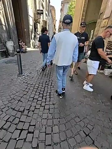 This video captures a bustling street scene in what appears to be Naples, Italy. The time of day is clearly daytime, indicated by the bright sunlight casting distinct shadows and the clear blue sky. The weather seems to be warm and pleasant, as many people are dressed in light clothing like t-shirts, shorts, and sandals.

The scene is a narrow, cobblestone street lined with tall, multi-story buildings. The architecture suggests an older, historic European city. The buildings have a mix of residential and commercial spaces, with shops and cafes visible. There are awnings and outdoor seating for cafes, adding to the lively atmosphere.

The primary subjects are the people walking along the street. They are a diverse group, including tourists and locals, engaged in everyday activities. Some are walking with purpose, while others are leisurely strolling and taking in the sights. One person in the foreground, walking away from the camera, is wearing a light blue shirt, a white vest, and jeans, carrying a book. To the right, another man in a black t-shirt and denim shorts is walking, holding a phone. Further down the street, more people are visible, some browsing at market stalls. There's a sense of vibrant activity and movement.

Notable details include the uneven cobblestone pavement, which is characteristic of many Italian cities. The sunlight plays a significant role, highlighting textures and creating a warm, inviting ambiance. The presence of a bell tower or a church steeple in the distance adds to the Italian context.

There are a few visible signs. One sign on a building reads "XENON". Another sign, partially visible on a stall, has illegible text.

The overall mood conveyed is one of a typical, lively day in a charming European city, with people enjoying the urban environment.