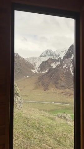 A video shows a view of mountains from inside a room. The camera zooms out from a window to reveal the landscape. The mountain range is partially snow-covered. A road with vehicle traffic is visible in the valley below. A grassy field sits in the foreground. The sky is overcast. The room has wood paneling.